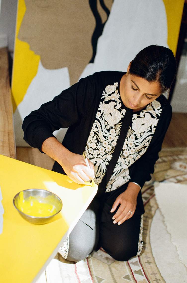 Rupy C. Tut kneels on a rug, painting the edge of a yellow surface with a brush; a bowl of yellow paint and a large artwork are nearby. he is wearing a matching black and floral set, and is an Indian-American woman with medium skin tone and long dark hair pulled back.