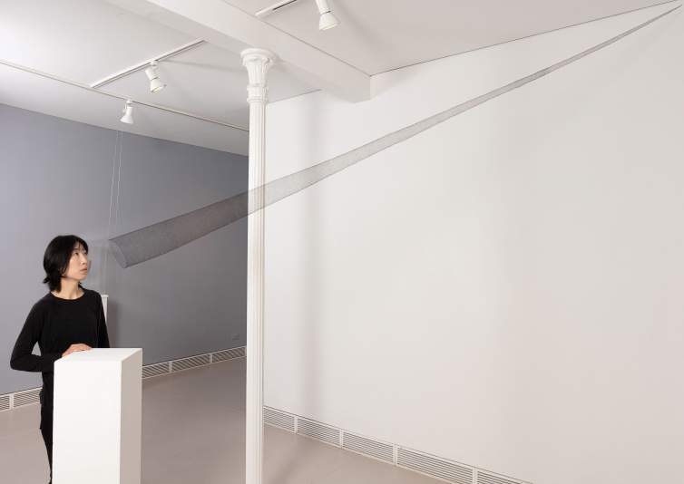 In a white gallery space, a person looks into sculpture “The Portal” which is a very elongated transparent cone made from very finely woven black hair that stretches from their face to the upper corner of the room.