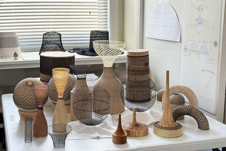 On a table in Jayoung Yoon’s studio, around a dozen wooden forms and translucent weavings sit together at varying heights and stages of completion. On one wall behind them is a window with slat blinds and woven forms sitting on the window sill. On the other wall, we see the artist’s sketches and templates for woven sculptures.