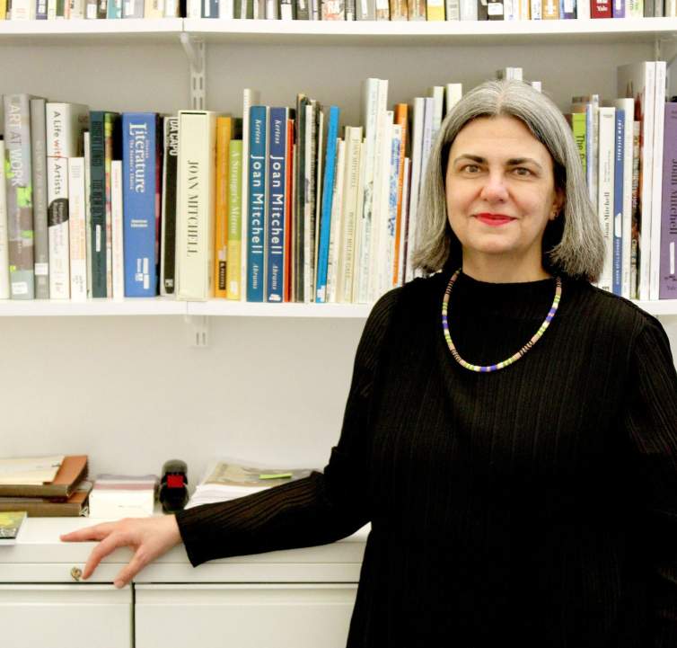 Alexandra Keiser stands in front of a bookshelf with various art catalogues. She is a white woman with shoulder length grey hair, wearing a black shirt and beaded necklace.