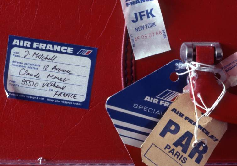A close up view of a red leather suitcase with Air France tags on it reading PAR Paris and JFK NY, and an address label with "J. MItchell, Vetheuil, France"