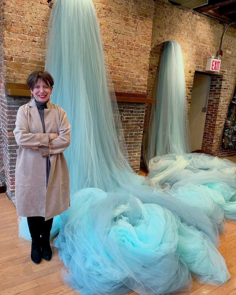 Ana María Hernando stands in front of two cascading waterfalls of blue tulle fabric in a brick walled room. She smiles with crossed arms, and is an Argentinian woman with dark short hair, wearing a trench coat and dark pants.