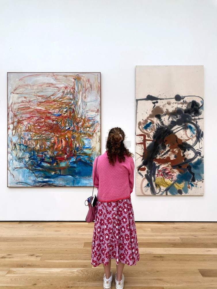 A woman with long curly hair stands in a gallery between two abstract paintings. The left painting has a tangle of swirling red and orange marks floating over a blue area, and the painting on the right has organic dark shapes punctuated by gestures of red, and blue splatter. She wears a pink sweater and patterned skirt.