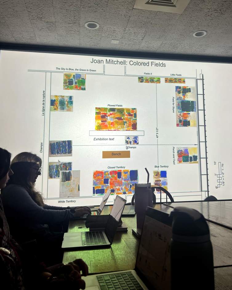 In a darkened lecture room, people sit around a central table with laptops looking at a screen showing a floor plan of an exhibition space with abstract paintings placed and labeled.
