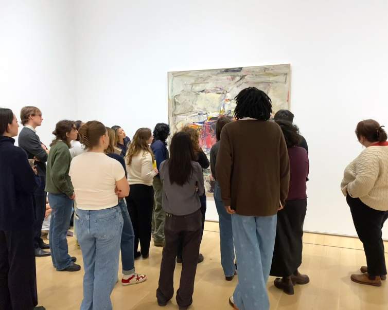 In a museum gallery space with white walls and wood floors, a group of 15 college students face a square abstract painting.