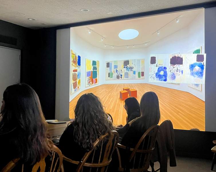 In a darkened lecture room, people sit around a central table looking at a screen showing a gallery space with multi-panel abstract paintings.
