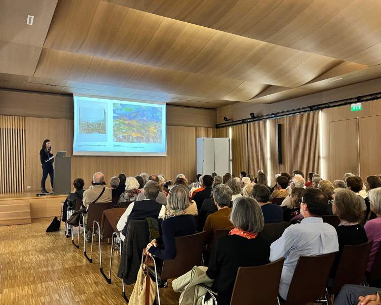 In a lecture hall with modern slat wood walls and ceilings, a white woman with long brown hair wearing a black top and pants presents a slide lecture to a seated crowd.
