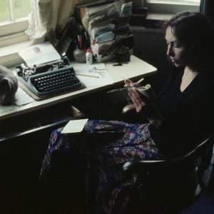 Carolee Schneemann sits in a wood armchair with one knee crossed over the other as she writes in a notebook, which appears to be long and narrow, like a reporter might use. She is mostly in shadow as sun falls through an open window onto a typewriter and other office supplies, and what might be edge of a cat loafing on the artist’s desk.