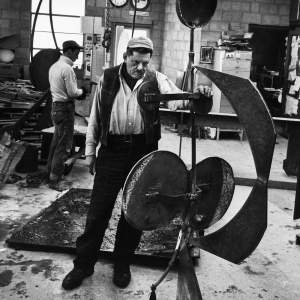 Black-and-white photograph of David Smith resting one hand on the crossbeam of a sculpture a full two heads taller than he is. He dips his chin and looks at the piece from under his eyebrows. The work is made up of disks, sections of arcs, and a cup-shaped form spaced along vertical support.