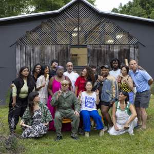 Fifteen people pose in two lines around a Mel Chin, who sits and holds up one index finger. Some of the people smile and some make faces. They are gathered on a grassy lawn in front of a building with wood paneling on its façade.