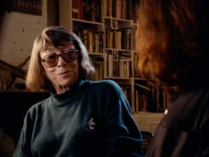 Joan Mitchell smiles in a darkened library room sitting for an interview. She is an older white woman with tinted glasses and brown hair in a blunt bob.