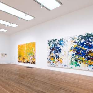 Several large abstract paintings hang in a white gallery space with several small framed prints, by artist Joan Mitchell.