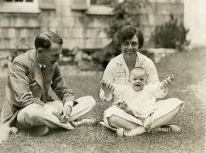 A white man and woman sit crosslegged in the grass, the woman smiles to camera and holds up the hands of a smiling baby