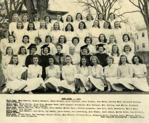 5 rows of young white women in white dresses, with 4 in black caps and gowns