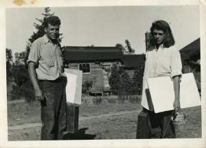 A young white man and white woman stand outside with serious expressions, each holding white drawing boards