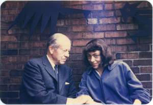 Joan Mitchell, a white woman with dark hair, smiles next to a white older man in a suit who looks to her and clasps her arm