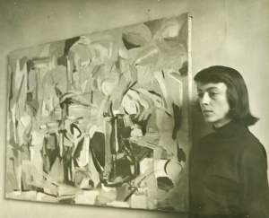 Joan Mitchell, a young white woman with dark hair, stands next to a cubist abstract painting gazing to the side