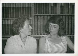 An older white woman turns to a younger white woman in front of a bookshelf
