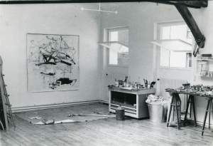 A square abstract painting hands on the wall, next to tables with art supplies and two open square windows