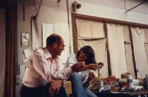 In a studio with canvas covering the windows, Joan Mitchell and Xavier Fourcade lean towards each other in conversation. She is a white woman with dark bob and glasses, and he is a white man with balding dark hair and glasses.