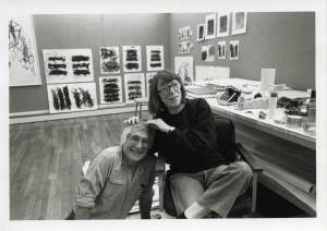In a studio lined with proofs of abstract prints on the wall, Joan Mitchell rests her hands on Ken Tyler's head, who smiles and crouches on the floor next to her chair.