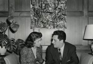 A white woman and man face each other on a couch below an abstract painting and wood panelled walls