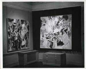 Two large square abstract paintings hang on dark gallery walls