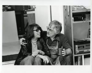 Joan Mitchell smiles in tinted glasses next to Ken Tyler whose arm is wrapped around her, each with a cigarette in hand.