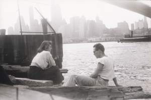 A white woman and  man in their 20s sit on the edge of the East River under the Brooklyn Bridge