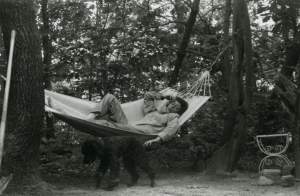 Joan Mitchell reclines in a white hammock in the woods, a black poodle stands near
