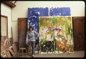 Artist Joan Mitchell leaning on a stool in front of 2 large paintings