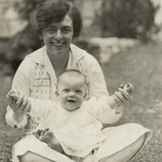A white woman with dark hair sits cross-legged in the grass, and holds up the hands of smiling baby all in white in her lap