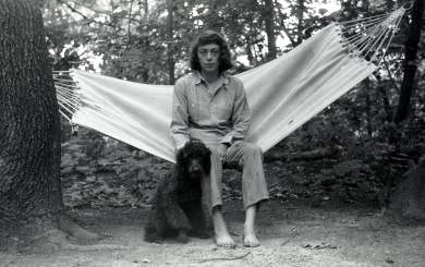 With dark hair cut in a bob, a white woman sits in a white canvas hammock among a grove of trees, with a black poodle sitting to the left of her bare feet.