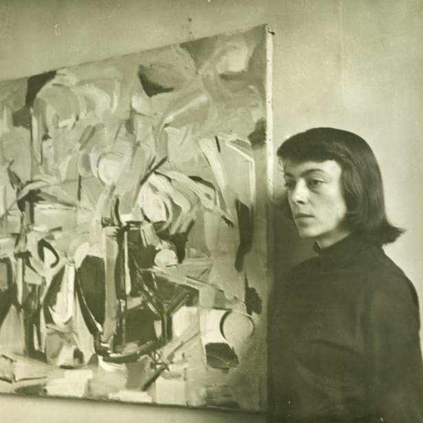 Joan Mitchell, a young white woman with dark hair, stands next to a cubist abstract painting gazing to the side