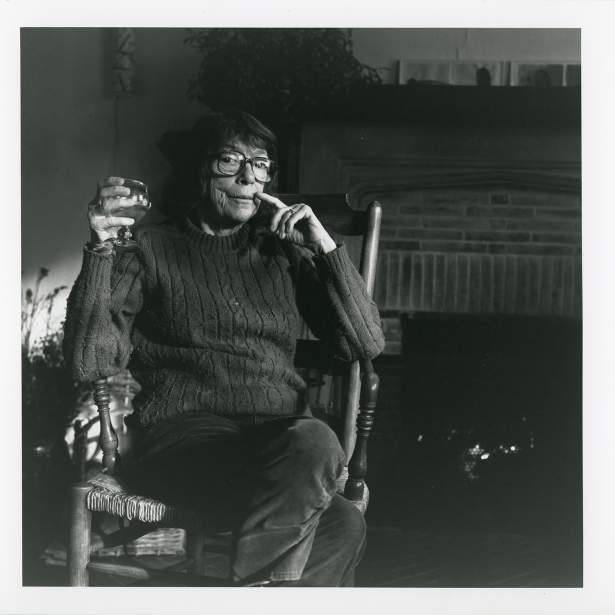 Joan Mitchell, an older white woman wearing large glasses and dark bobbed hair, sits in a chair with a hand to her face and holding a drink with a serious expression.