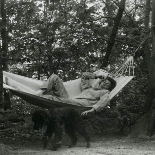 Joan Mitchell reclines in a white hammock in the woods, a black poodle stands near
