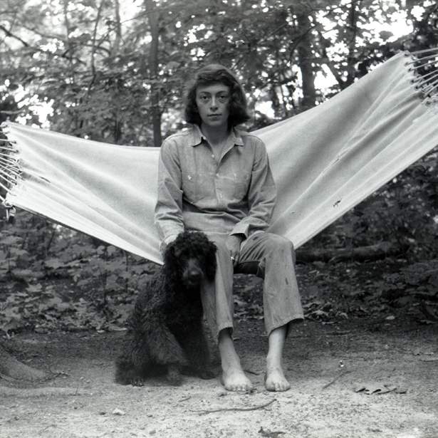 With dark hair cut in a bob, a white woman sits in a white canvas hammock among a grove of trees, with a black poodle sitting to the left of her bare feet.