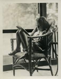A young white girl curled up in a rattan chair reading a book