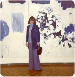 Joan Mitchell stands in front of her large blue and white abstract painting. She is a white woman in her 50s with a short dark bob haircut and is wearing all blue.