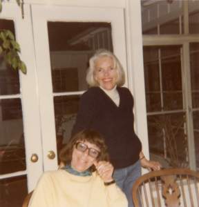Sally Perry, a white woman with white hair, smiles and stands above a sitting and smiling Joan Mitchell, a white woman with a dark bob.