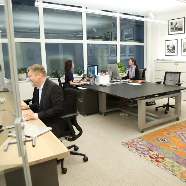 3 smiling staff members sit at desks in a well lit office room, with photographs of Joan Mitchell on back wall