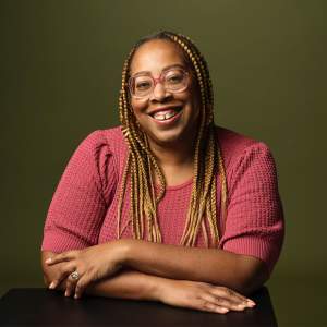 Angela Tucker smiles warmly with arms folded in front of her. She is a Black woman with a medium skintone, long honey blond-brown braids, and wears round fuschia glasses and matching knit shirt.