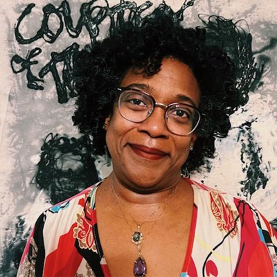 Cheryl Hayes smiles in front of a painting with abstract gestures and scribbled letters. She has medium dark skin and curly black hair, and is wearing wire glasses, a brightly patterned dress, and two pendant necklaces.