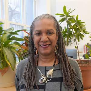 Michele Brierre smiles warmly in an office filled with plants. She is , a Black woman medium skin tone and long gray braids, wearing a silver necklace and geometric patterned sweater.
