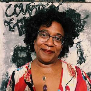 Cheryl Hayes smiles in front of a painting with abstract gestures and scribbled letters. She has medium dark skin and curly black hair, and is wearing wire glasses, a brightly patterned dress, and two pendant necklaces.