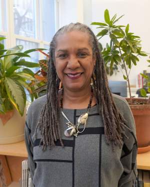Michele Brierre smiles warmly in an office filled with plants. She is , a Black woman medium skin tone and long gray braids, wearing a silver necklace and geometric patterned sweater.