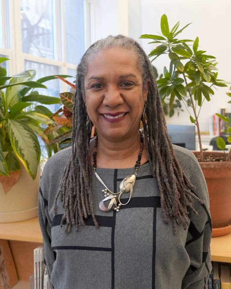 Michele Brierre smiles warmly in an office filled with plants. She is , a Black woman medium skin tone and long gray braids, wearing a silver necklace and geometric patterned sweater.