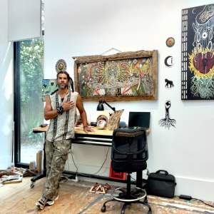 Mankh stands with a hand over his heart in front of a work table, with various abstract artworks hanging on the wall in a white walled studio with a tall window and paint-splattered floor. He is an Afro-indigenous man with medium skin tone, long dark dreadlocks pulled back, and a beard, wearing patterned sleeveless shirt and camouflage pattern pants.