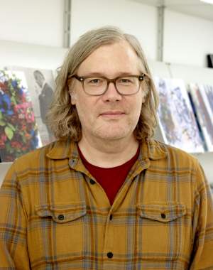 Andrew Beccone, a white man with shoulder length blond hair wears glasses and a plaid shirt.
