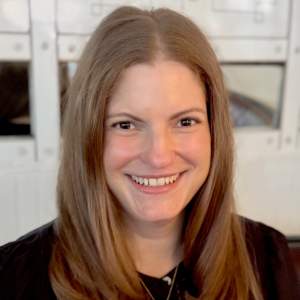 Christina de Gersdorff smiles warmly in front of a white wall with frames. She is a white woman with light skin tone, long light brown hair, and brown eyes.